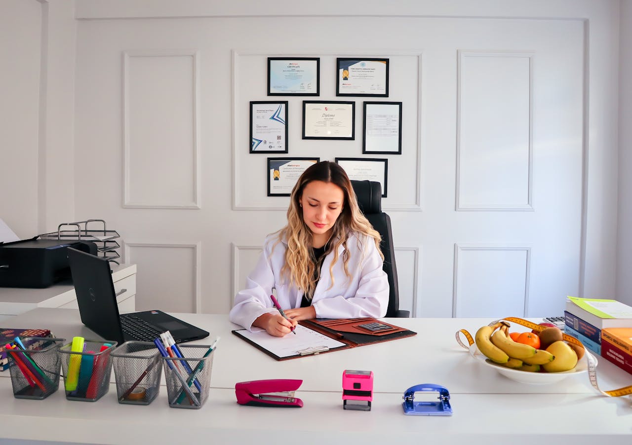 Nutritionist sits at a modern office desk writing, with fruits and diplomas displayed, emphasizing health and professionalism.